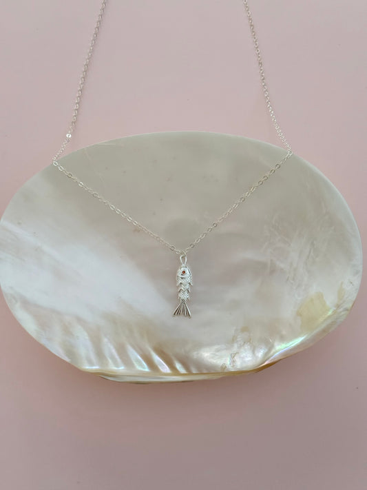 A delicate silver fish pendant necklace displayed on a large, iridescent white shell dish against a soft pink background.