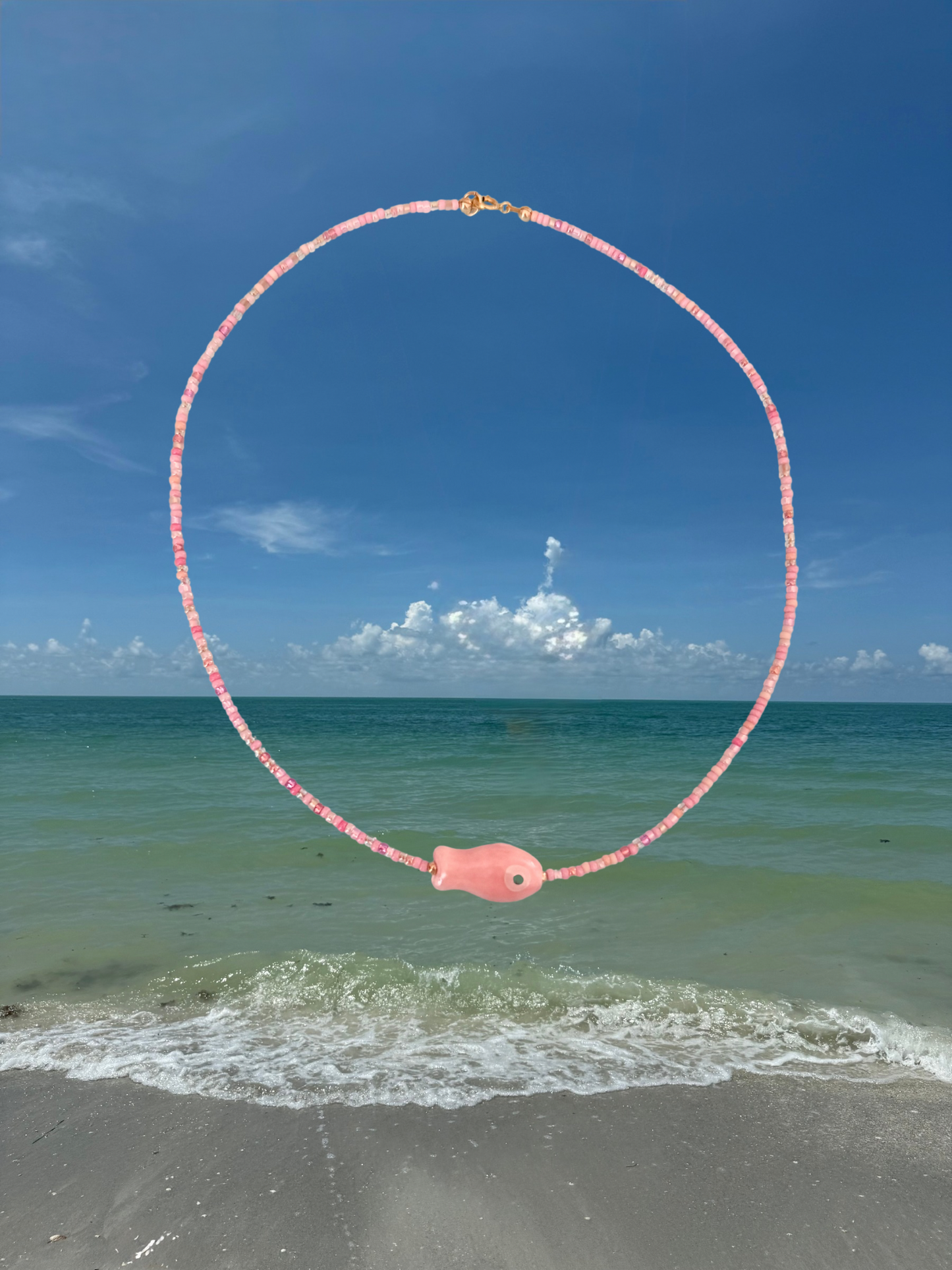 Necklace with pink fish pendant over a beach and ocean scene