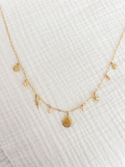 A gold filled charm necklace with a dainty paper clip chain and multiple small charms including a shell and other sea-themed shapes, displayed on a white textured surface.