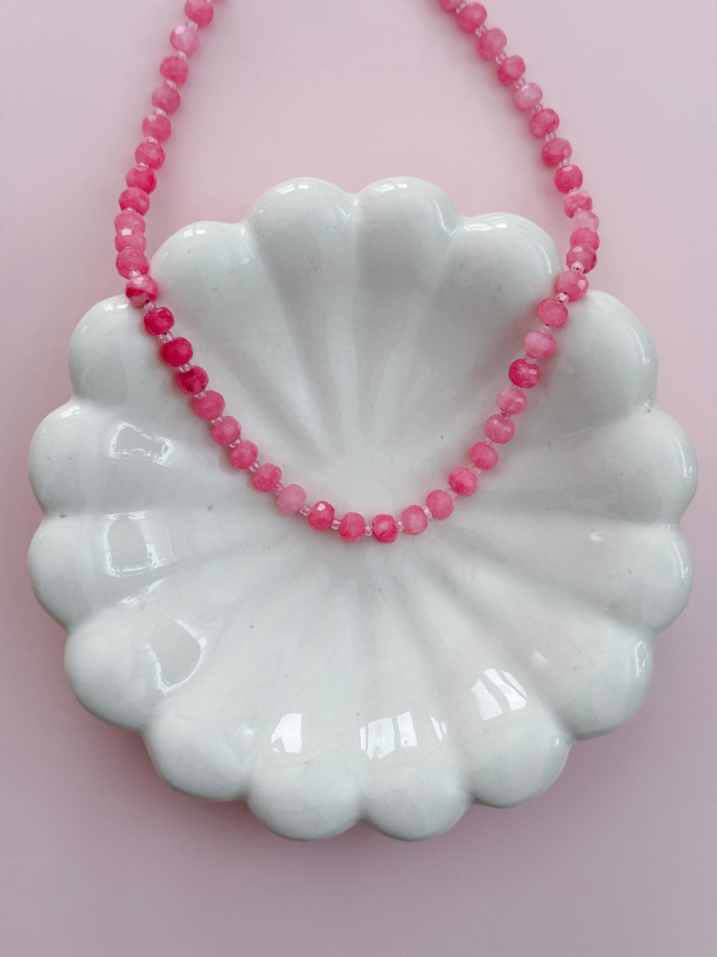 A pink beaded necklace draped over a white scalloped ceramic dish against a soft pink background.