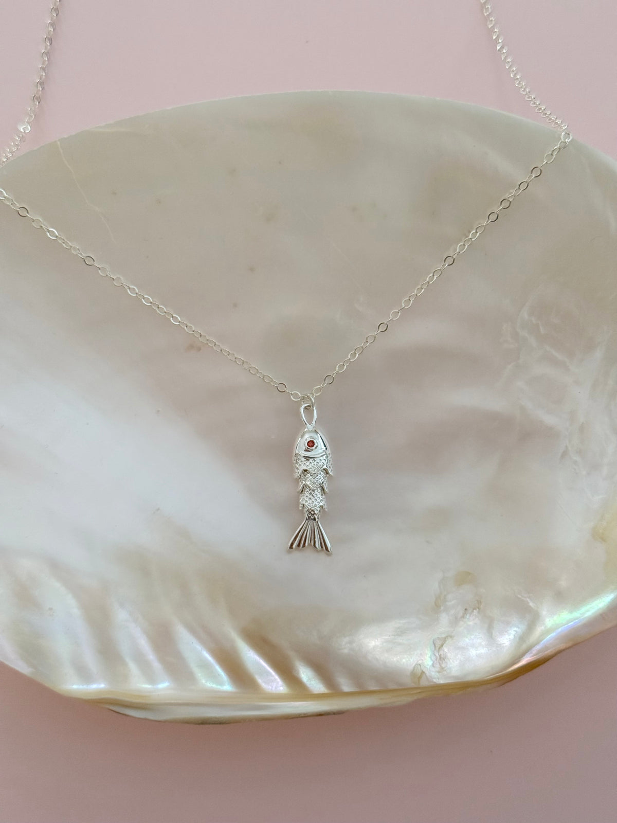 A delicate silver fish pendant necklace resting on a large, iridescent mother-of-pearl shell against a soft pink background.