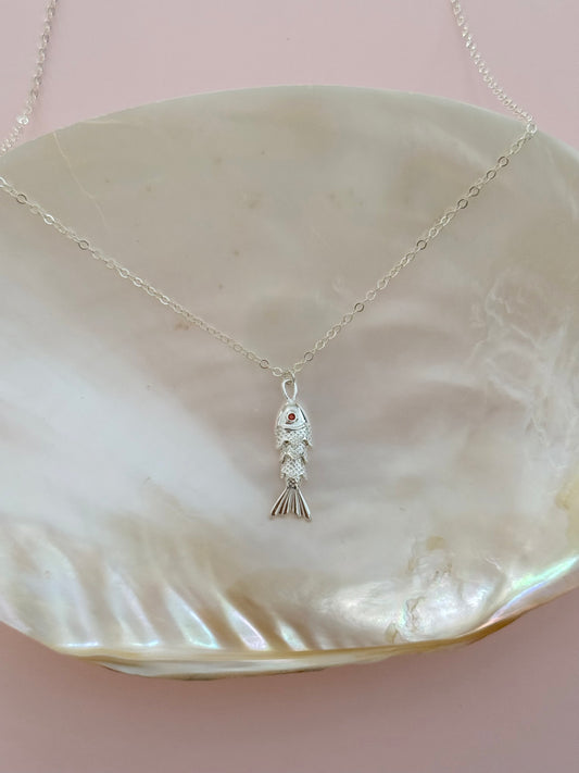 A delicate silver fish pendant necklace resting on a large, iridescent mother-of-pearl shell against a soft pink background.