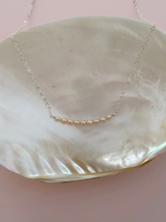 A delicate silver chain necklace with a small curved row of pale pearl-like beads, displayed on a large iridescent shell dish against a soft pink background.