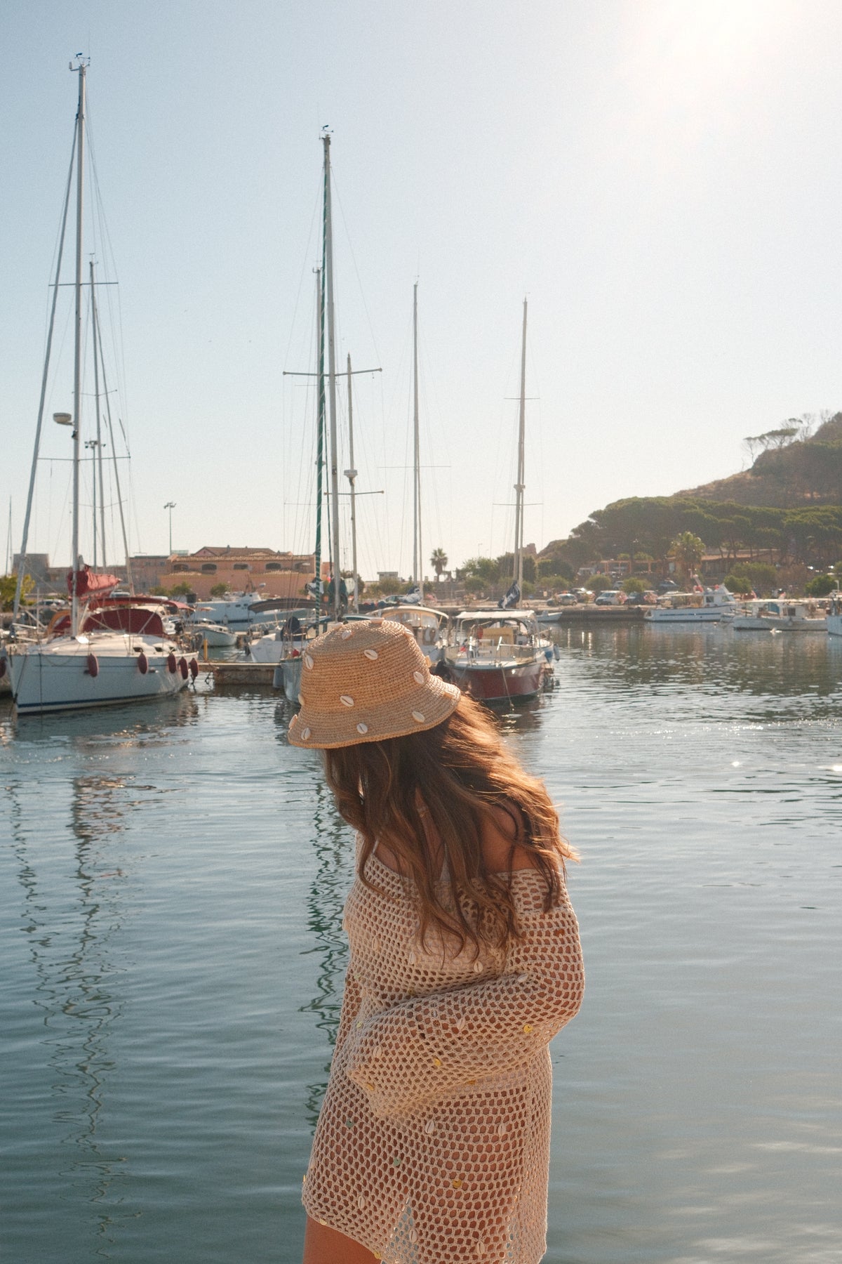 A woman wearing a beige crochet top and a matching bucket hat stands by the water at a harbor, with boats and a shoreline visible in the background.