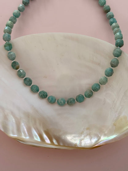A beaded necklace made of faceted round beads in a greenish turquoise-blue hue, displayed on a large iridescent shell against a soft pink background.