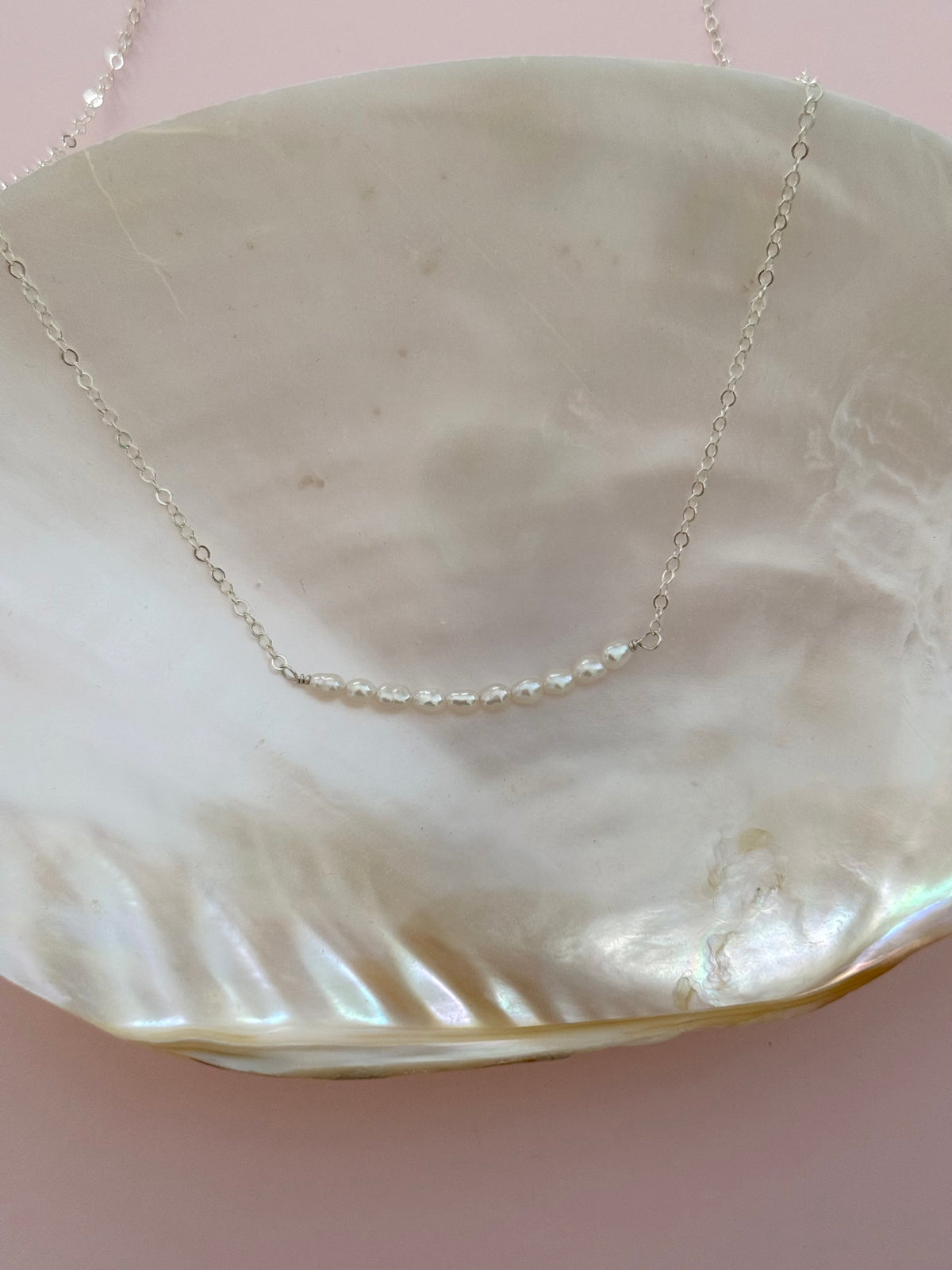A delicate silver chain necklace with a short segment of small white pearls, displayed on a large iridescent shell against a soft pink background.