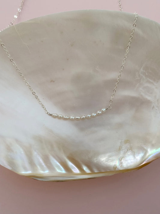 A delicate silver chain necklace with a short segment of small white pearls, displayed on a large iridescent shell against a soft pink background.