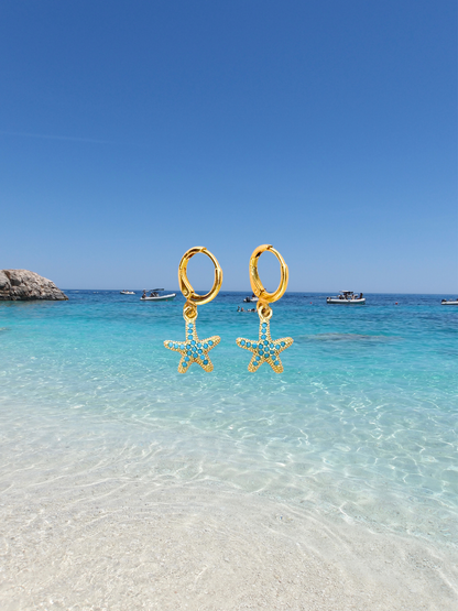 A pair of gold hoop earrings with dangling starfish charms embellished with small light blue stones, shown against a tropical beach backdrop with turquoise water and a clear blue sky.
