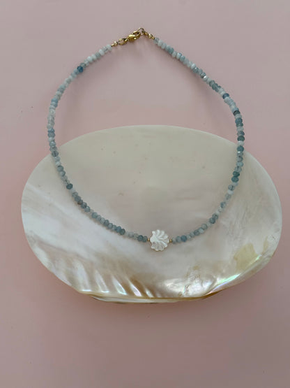 A delicate beaded necklace with light blue-gray round beads, a small white shell flower charm, and a gold clasp, displayed on a white mother-of-pearl shell dish against a soft pink background.