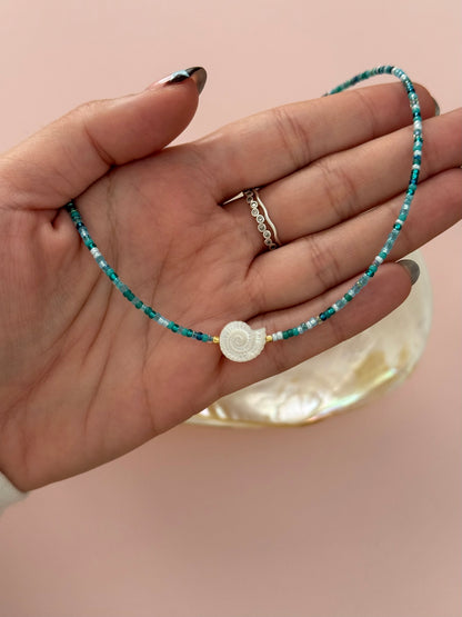 A hand holding a delicate beaded necklace with turquoise and white beads, featuring a central white spiral shell charm, set against a soft pink background.