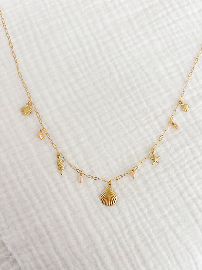A gold filled charm necklace with a dainty paper clip chain and multiple small charms including a shell and other sea-themed shapes, displayed on a white textured surface.
