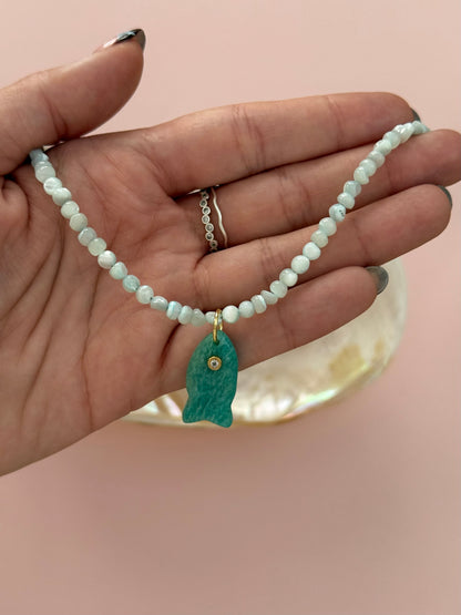 A hand holding a beaded necklace with a small turquoise fish pendant and a gold accent, set against a soft pink background.