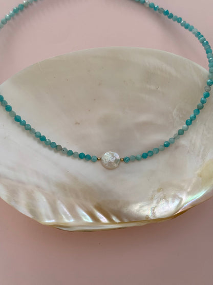 A beaded necklace with turquoise-blue faceted beads and a central white shell bead, displayed on a large white shell against a soft pink background.