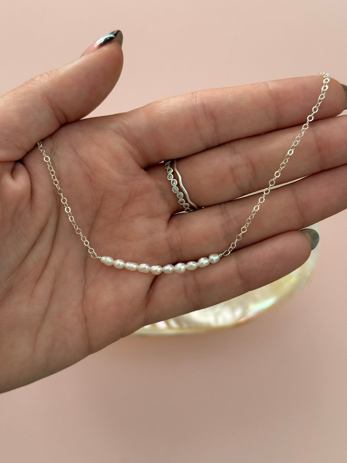 A delicate silver chain necklace with a short segment of small white pearls, held in a person's hand against a soft pink background.