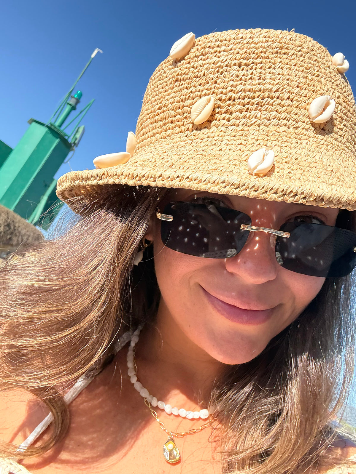 A close-up lifestyle shot of a woman wearing a beige woven straw hat adorned with cowrie shells, black sunglasses, and a pearl necklace against a blue sky background.