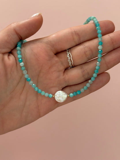 A beaded bracelet with turquoise and white beads, featuring a central white oval stone, displayed on a person's hand against a solid pink background.