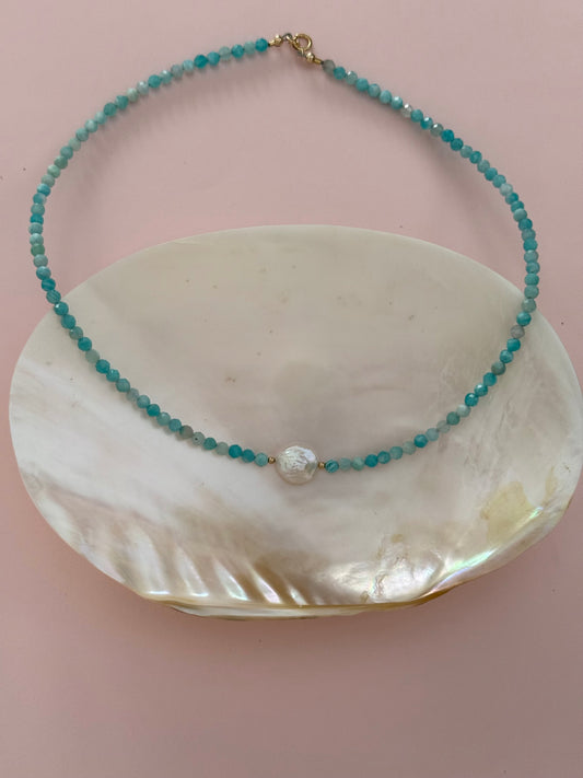 A beaded necklace with turquoise-blue faceted beads, a central white pearl-like bead, and a gold clasp, displayed on a white shell dish against a soft pink background.