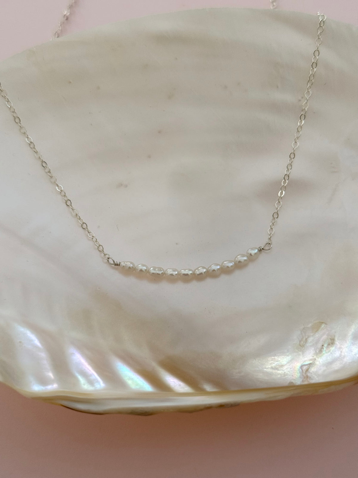 A delicate silver chain necklace with a short segment of clear faceted beads, displayed on a large iridescent white clam shell against a soft pink background.