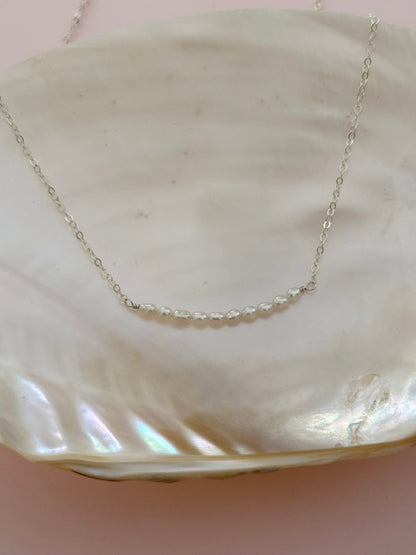 A delicate silver chain necklace with a short segment of clear faceted beads, displayed on a large iridescent white clam shell against a soft pink background.