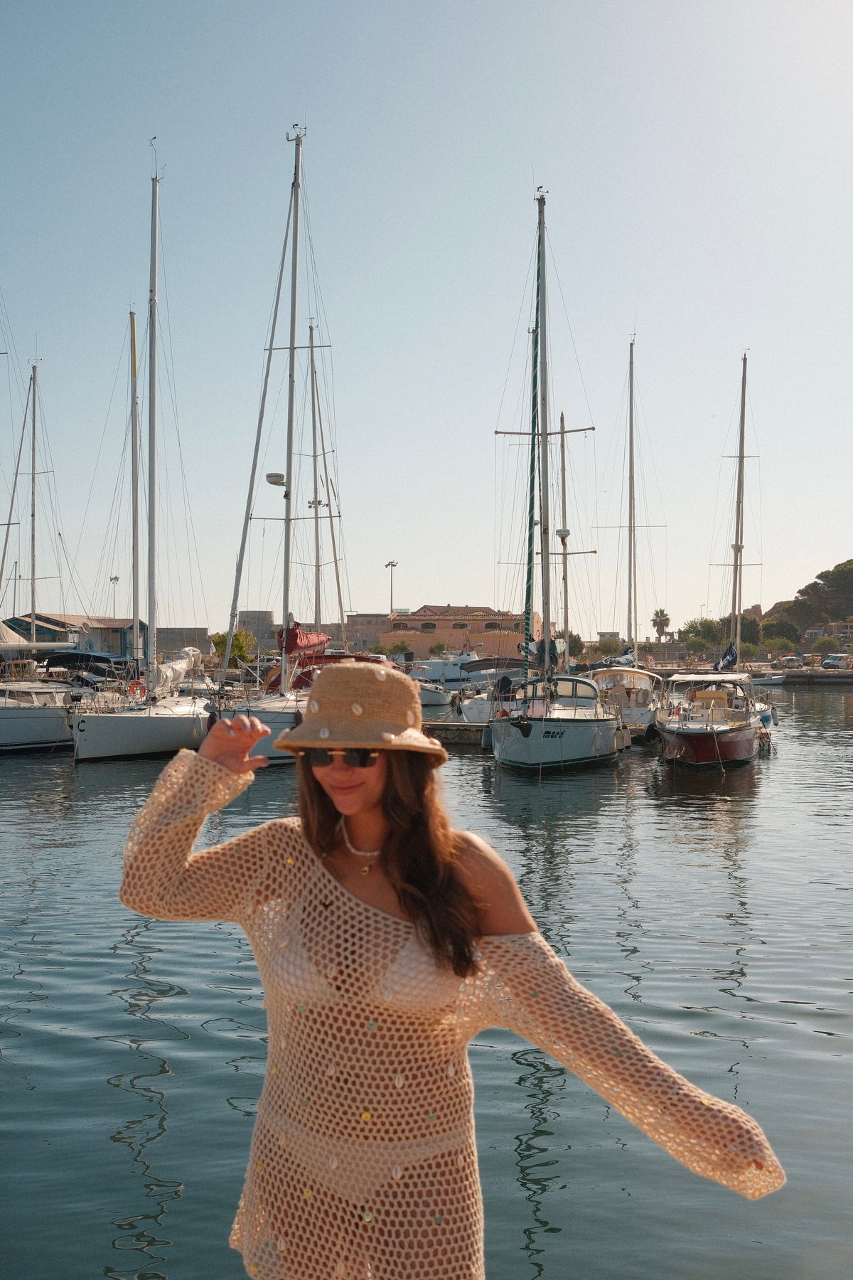 A woman wearing a beige crochet knit long-sleeve dress and a matching straw hat stands in a marina with boats in the background.
