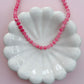 A pink beaded necklace draped over a white scalloped ceramic dish against a soft pink background.