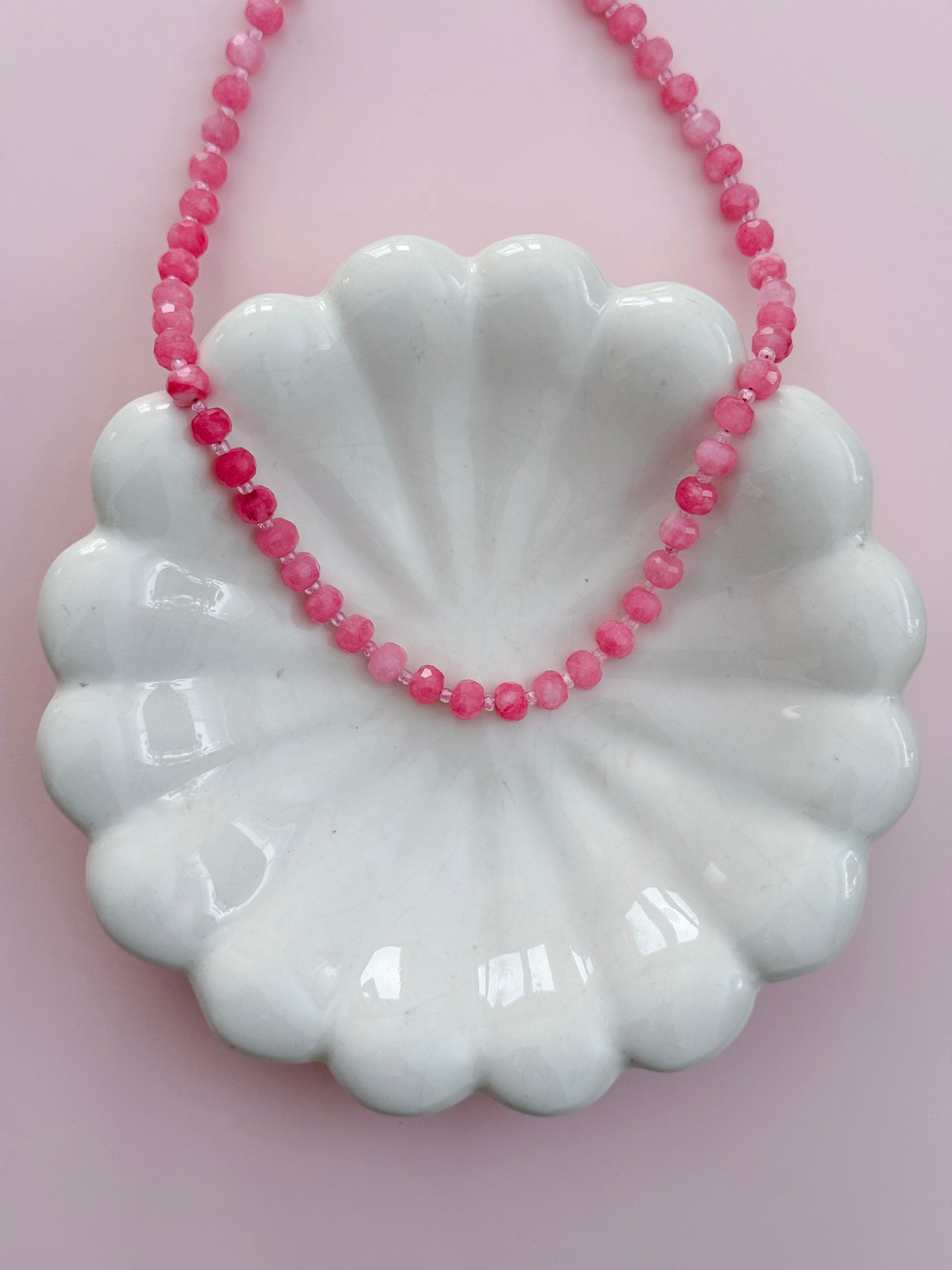 A pink beaded necklace draped over a white scalloped ceramic dish against a soft pink background.