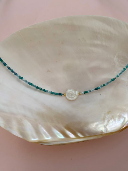 A delicate beaded necklace with teal and white beads and a spiral shell pendant, displayed on a large white clam shell against a soft pink background.