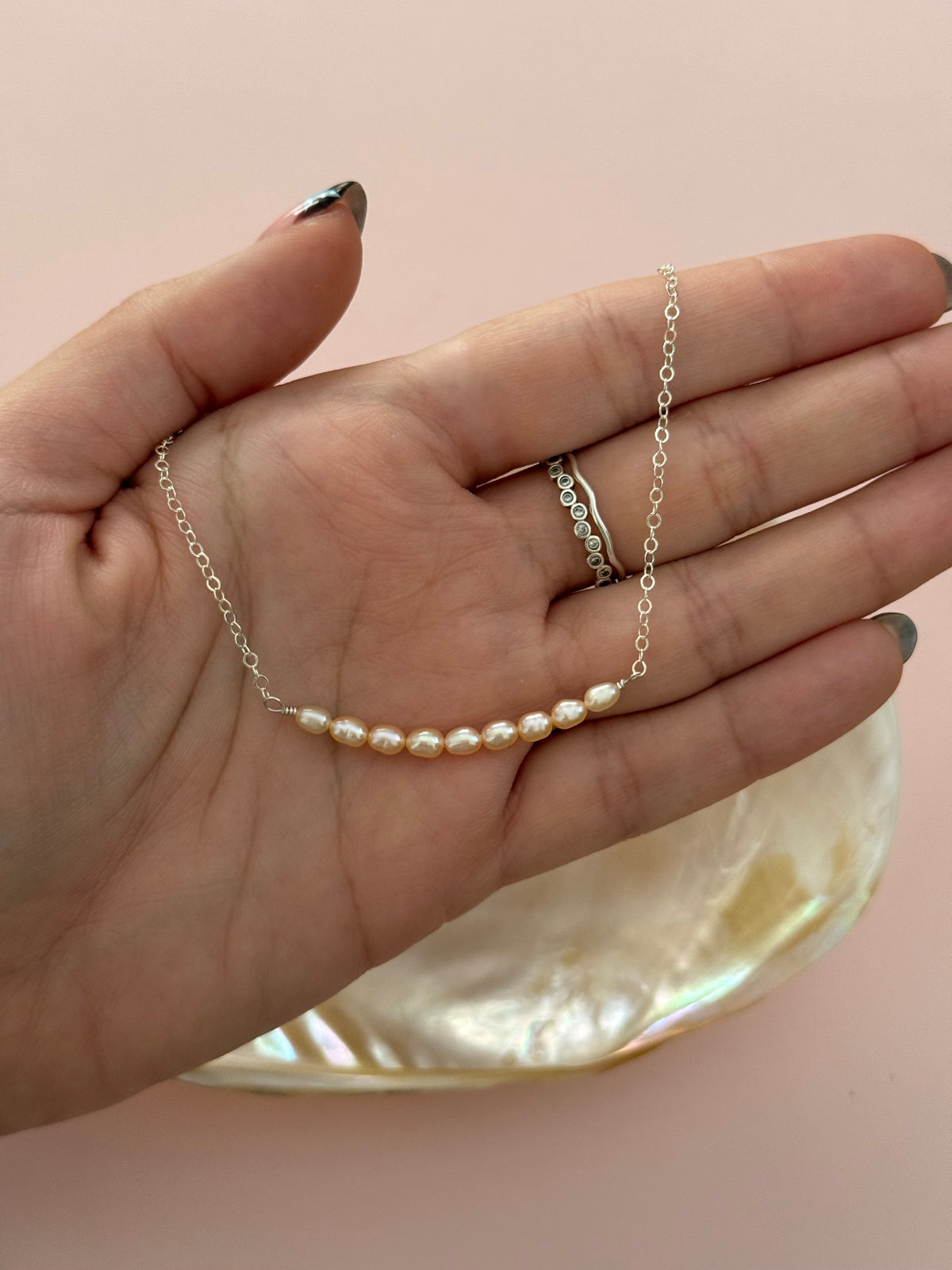 A delicate silver chain necklace with a curved row of small white freshwater pearls, held in a person's hand over a pink background with a mother-of-pearl dish.