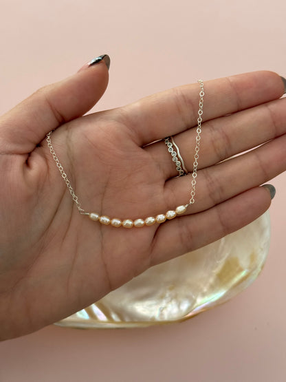A delicate silver chain necklace with a curved row of small white freshwater pearls, held in a person's hand over a pink background with a mother-of-pearl dish.