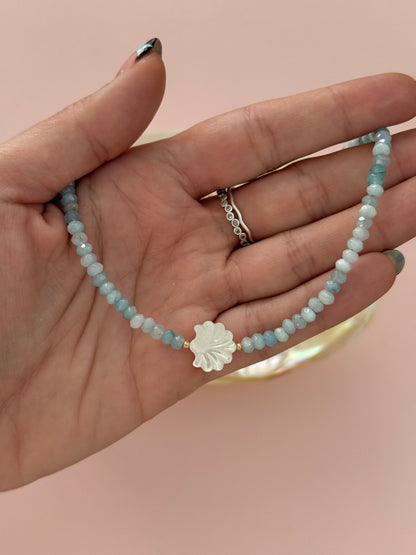 A close-up of a hand holding a beaded bracelet made of light blue and white round beads with a white flower-shaped charm in the center. The hand is shown against a soft pink background.